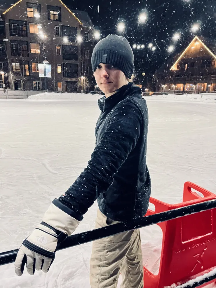 Ruby in a beanie and gloves at the rail of an outdoor ice rink while it snows