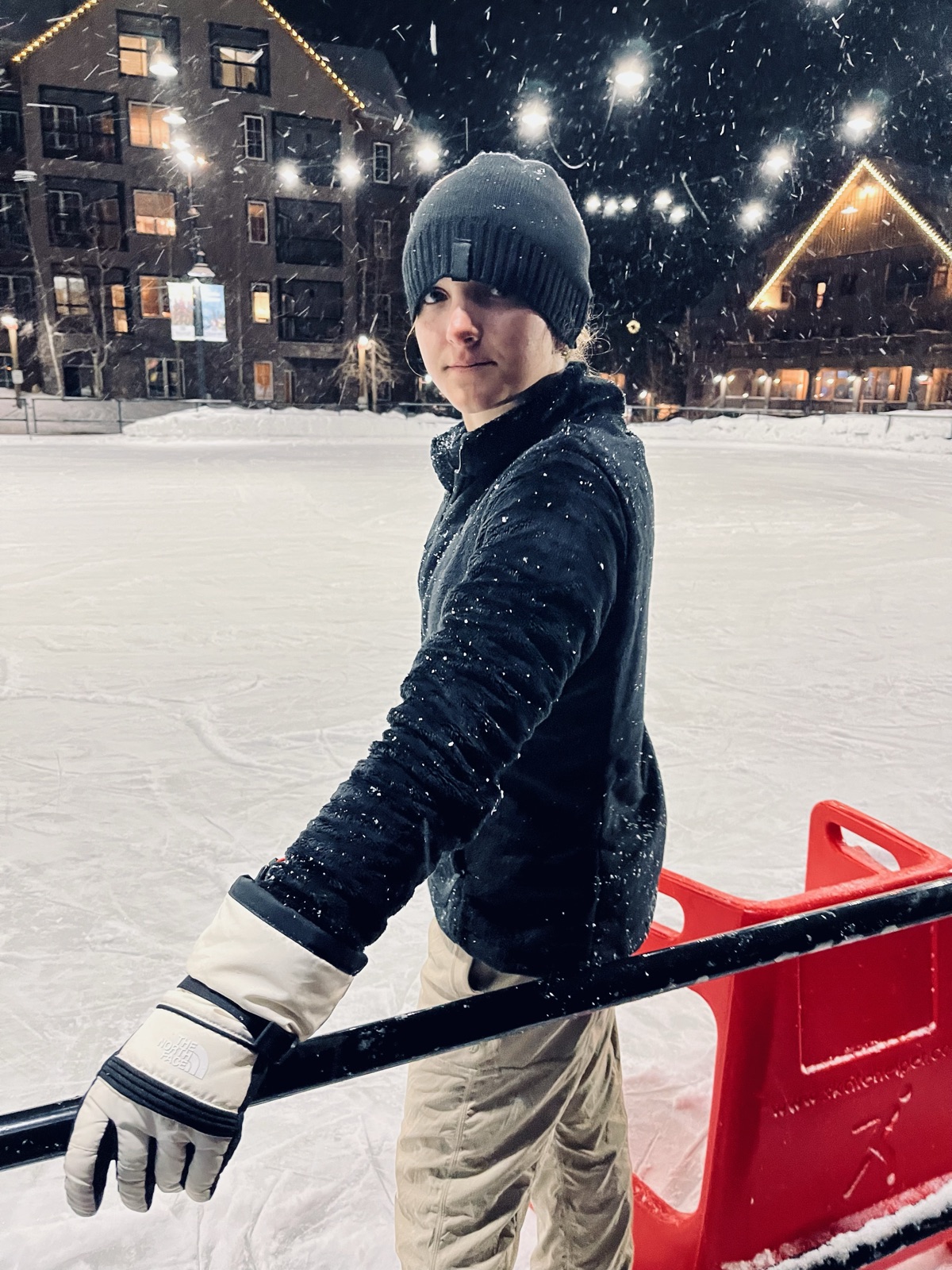 Ruby in a beanie and gloves at the rail of an outdoor ice rink while it snows