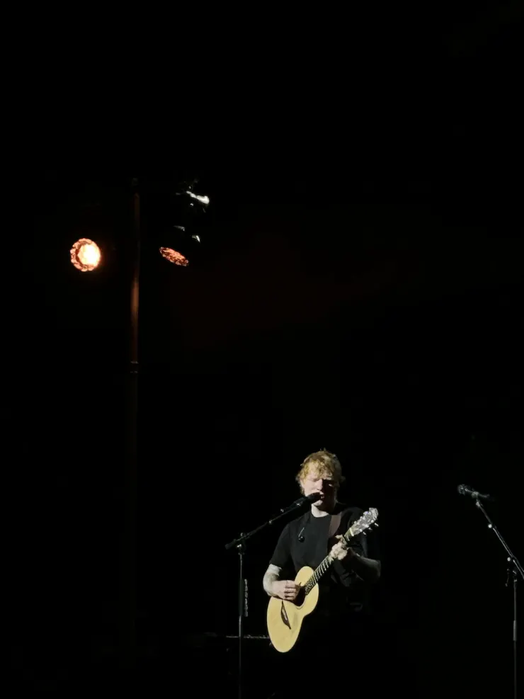 Ed Sheeran alone in a spotlight on a dark stage with his guitar