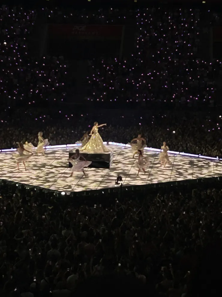 Dancers on a bright stage surrounded by a dark stadium full of phone lights