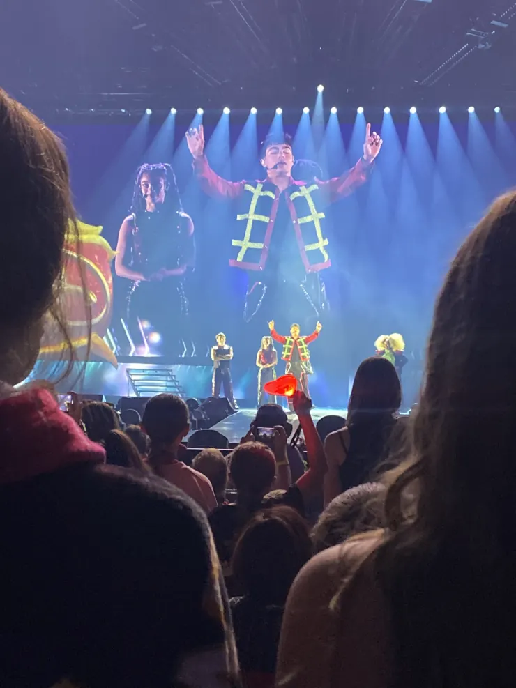 A performer on stage in a lit costume, arms raised, seen over the heads of the crowd