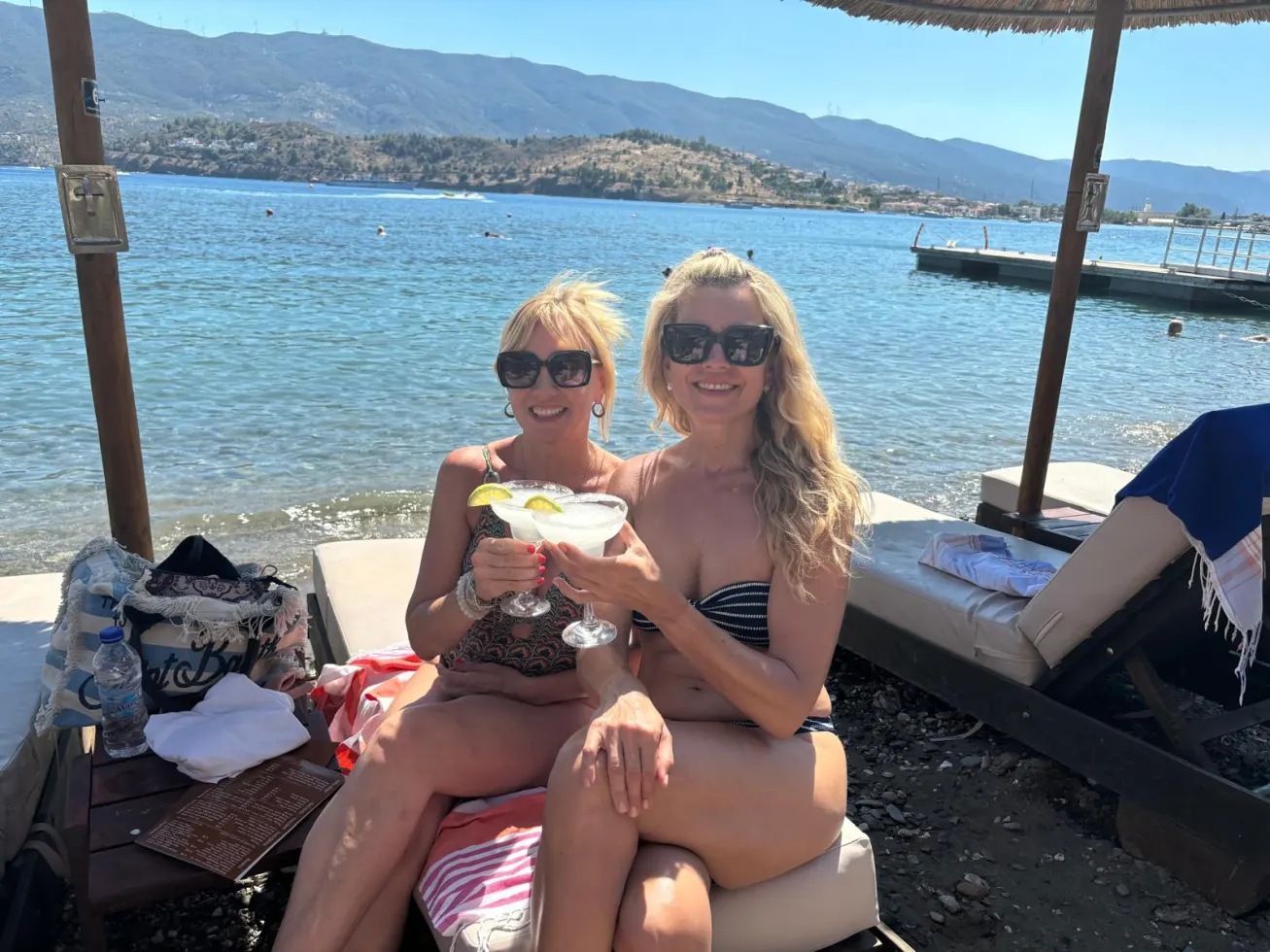 Two women toasting margaritas at a beach club, sea and hills behind