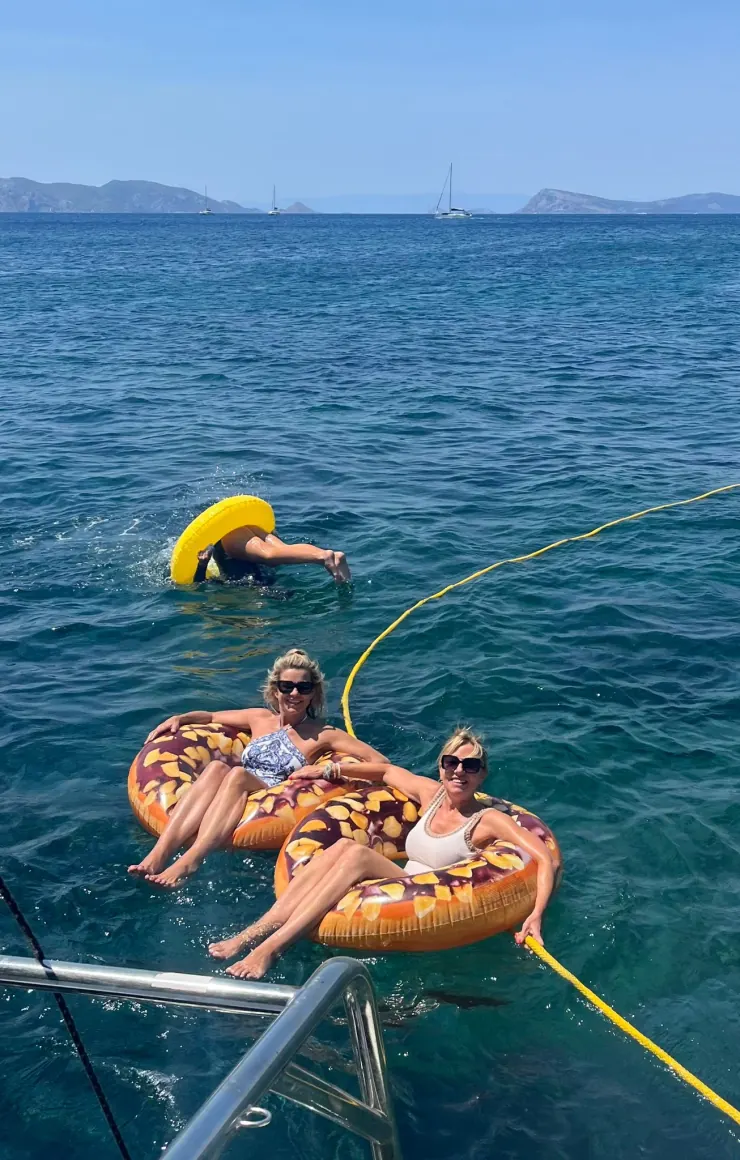 Two women floating on donut rafts beside a yacht, a swimmer nearby