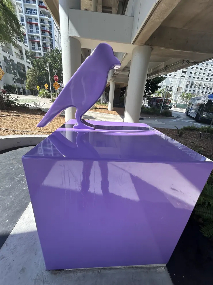 A purple bird sculpture on a plinth under a Miami overpass