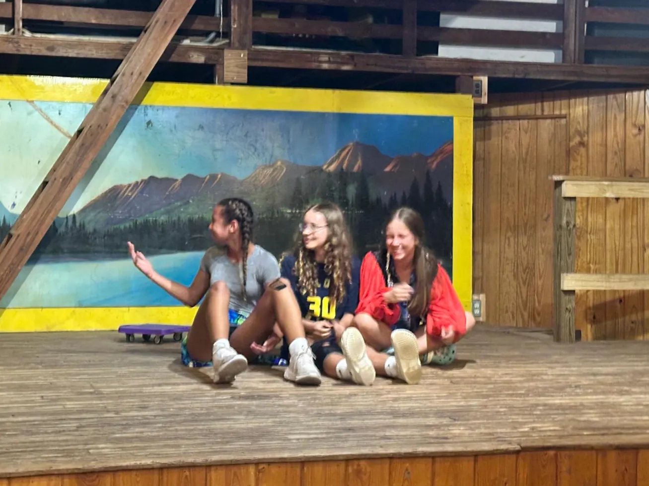 Three girls sitting cross-legged on a stage in front of a painted mountain backdrop