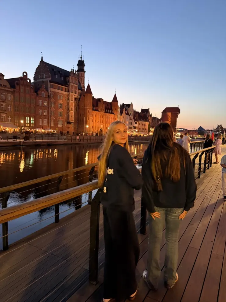 Ruby and a friend on a riverside boardwalk at golden hour, Gdańsk's old town behind them